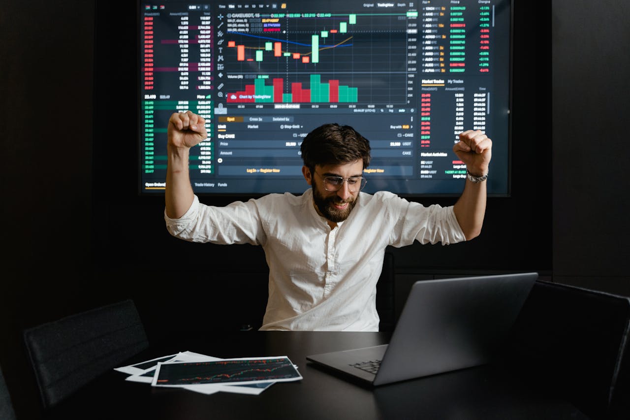 gallery-1 Businessman celebrates stock market success with hands raised in excitement at a trading desk.